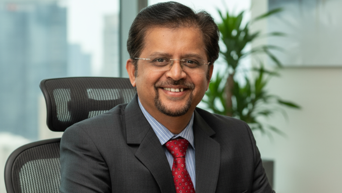 A professional portrait of Karthikeyan Natarajan, Co-CEO of Infinite Uptime. The background is a bright, soft-focus office setting with a green potted plant and large windows overlooking a city skyline.