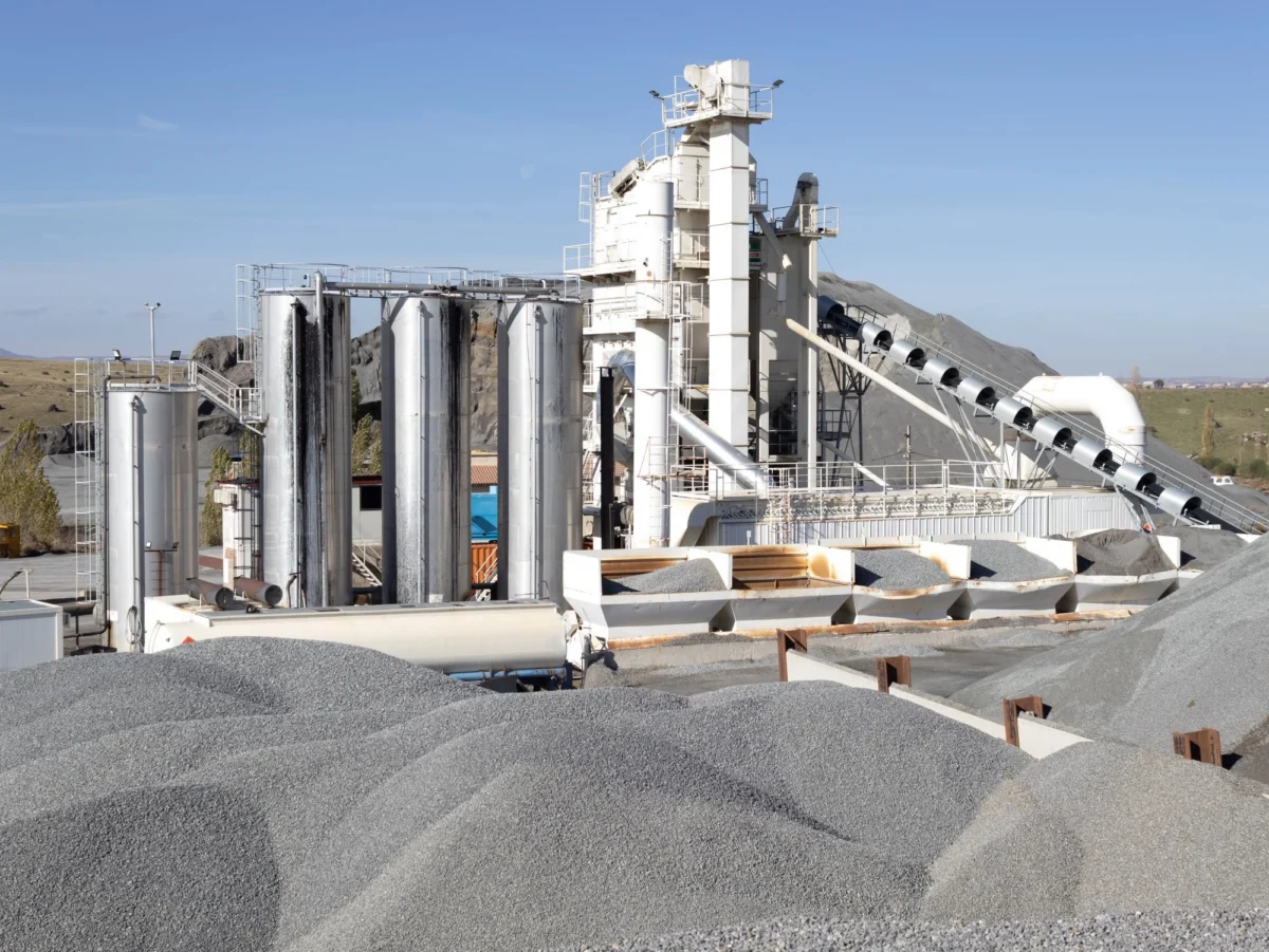 A large industrial processing site with four tall, cylindrical silver silos on the left and a central white multi-story machinery tower. A long diagonal conveyor belt extends to the right. In the foreground and background, there are massive, undulating mounds of grey crushed gravel under a bright, clear sky.