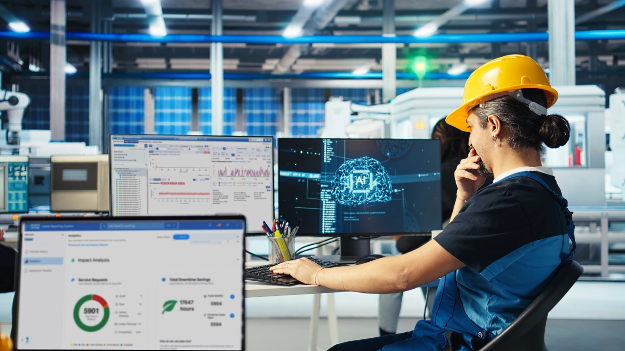 A woman in a hard hat and work overalls sits at a desk in a factory setting, working on multiple computer screens displaying graphs, charts, and data related to production and AI 4 Production Outcomes. One screen shows a brain-like graphic with "AI" prominently displayed, while industrial equipment is visible in the well-lit factory background.